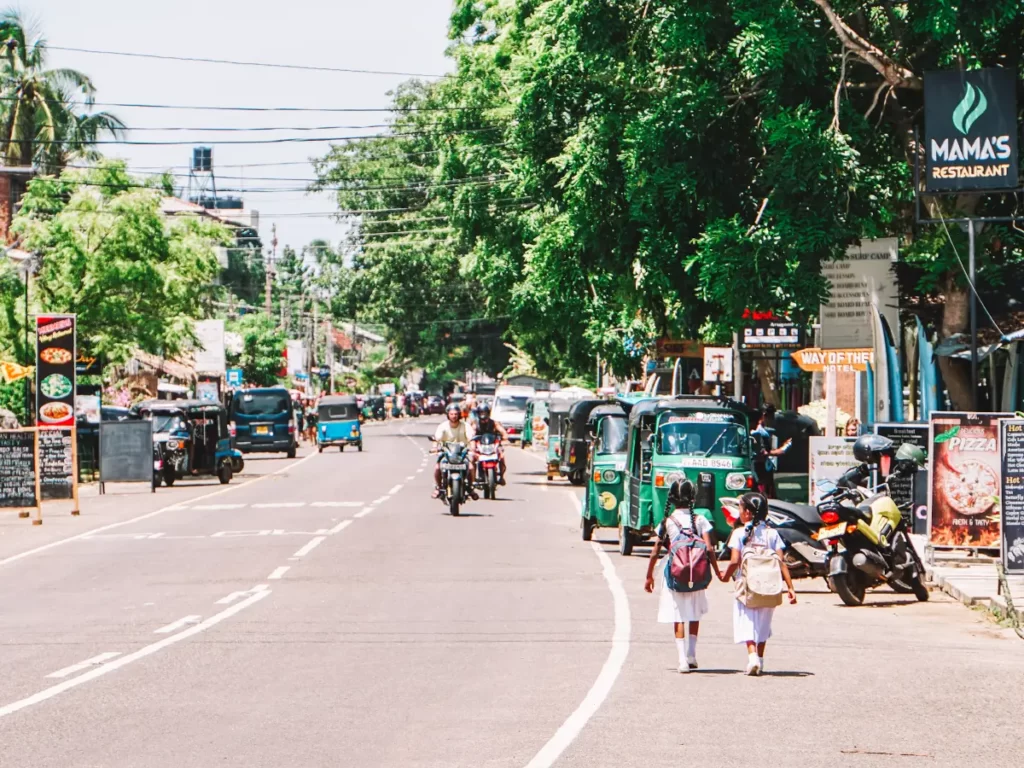 Two school girls hold hands as they walk down Arugam Bay main street with motorbikes driving past