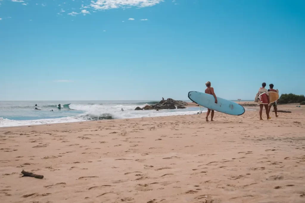 Three girls carry surfboards along the beach at Peanut Farm with surfers on a wave in the background