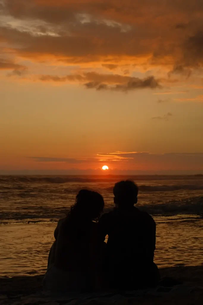 A silhouette of a couple sitting on Madiha Beach at sunset with a small red sun in the distance 