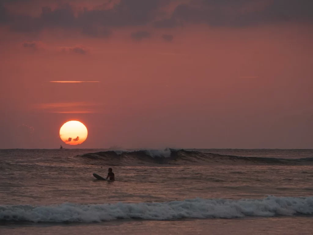 A person sits on a surfboard whilst a wave breaks in the background at The Rock Kabalana at sunset