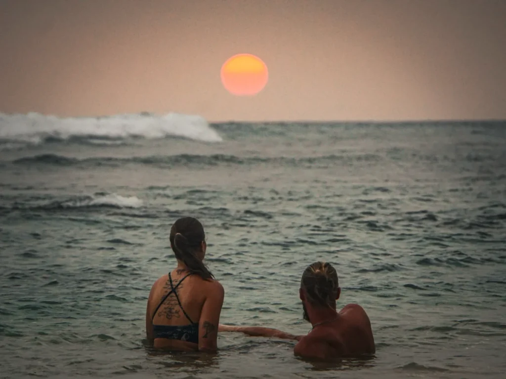 A couple sitting in the ocean with a low orange sun at the horizon near Ahangama Beach at sunset