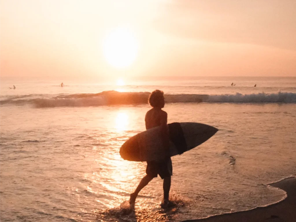 A man walks along Arugam Bay beach with a surfboard at sunrise